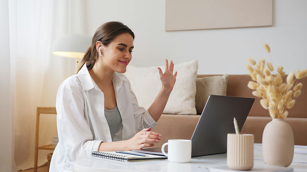 Woman wearing earbuds waves at laptop, smiling in cozy living room. Beige tones, vase with flowers, notebook, and mug on table.