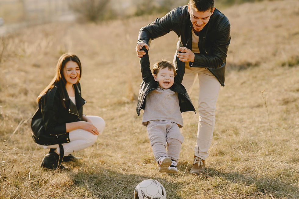 Family playing in a sunny field. A man holds a laughing toddler's hands, while a woman crouches nearby. Ball on grass, joyful mood.