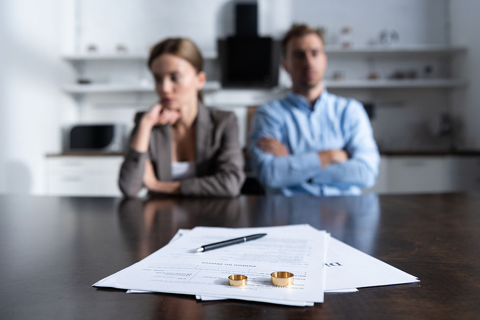A couple sits with arms crossed behind divorce papers and rings on a table in a bright office. The mood is tense and somber.