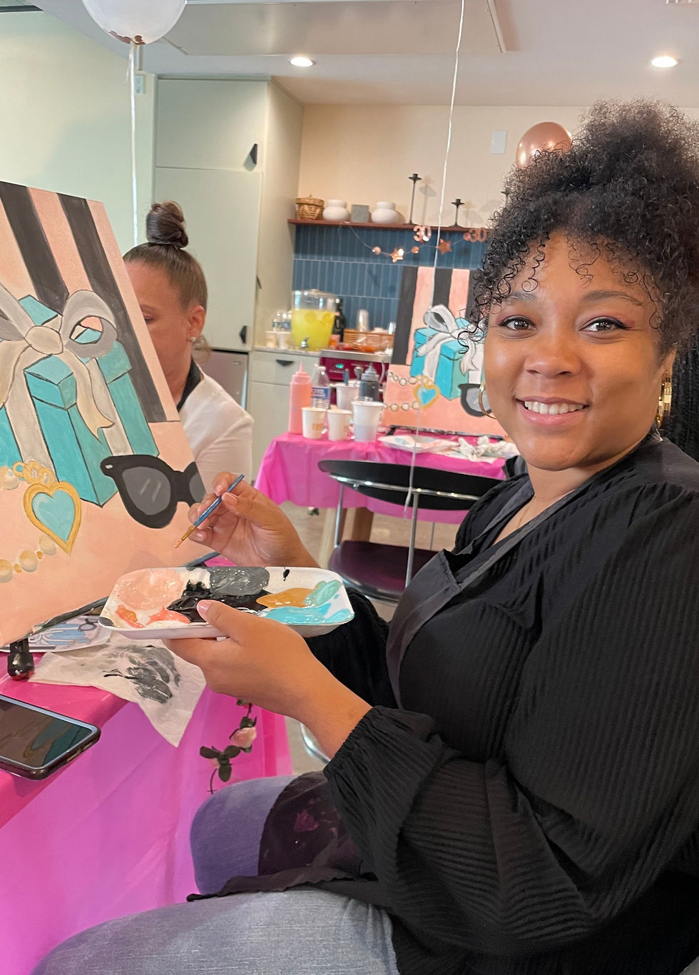 smiling lady holding paint palette and brush. She is sitting in front of her painting of a blue box and sunglasses.
