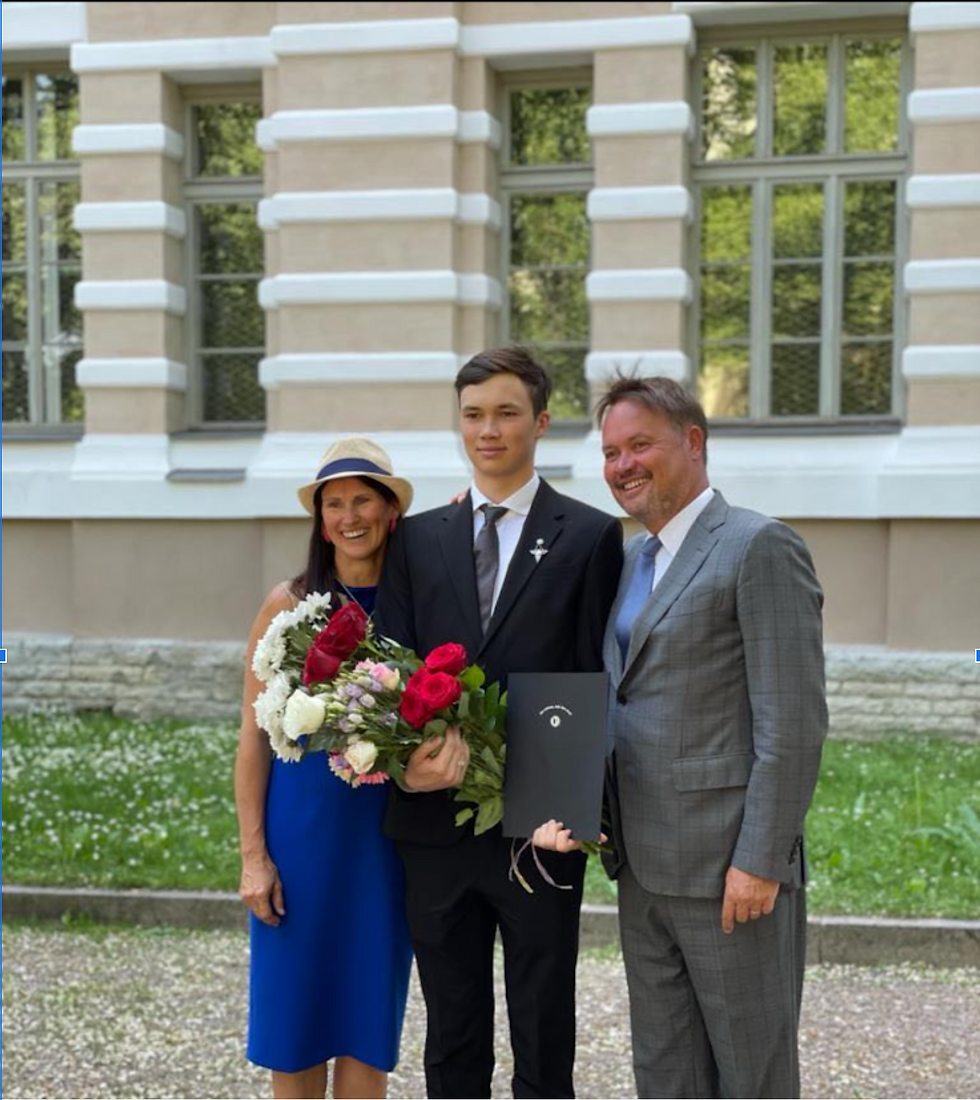 Young man in a suit holds flowers and a diploma, standing between a woman in a blue dress and a man in a gray suit, smiling outdoors.
