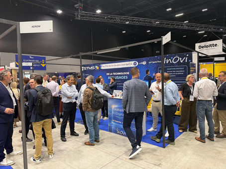 Salon pro bondé, hommes en discussion devant les stands STMS, Inovi et Deloitte; panneaux bleus, ambiance studieuse.
