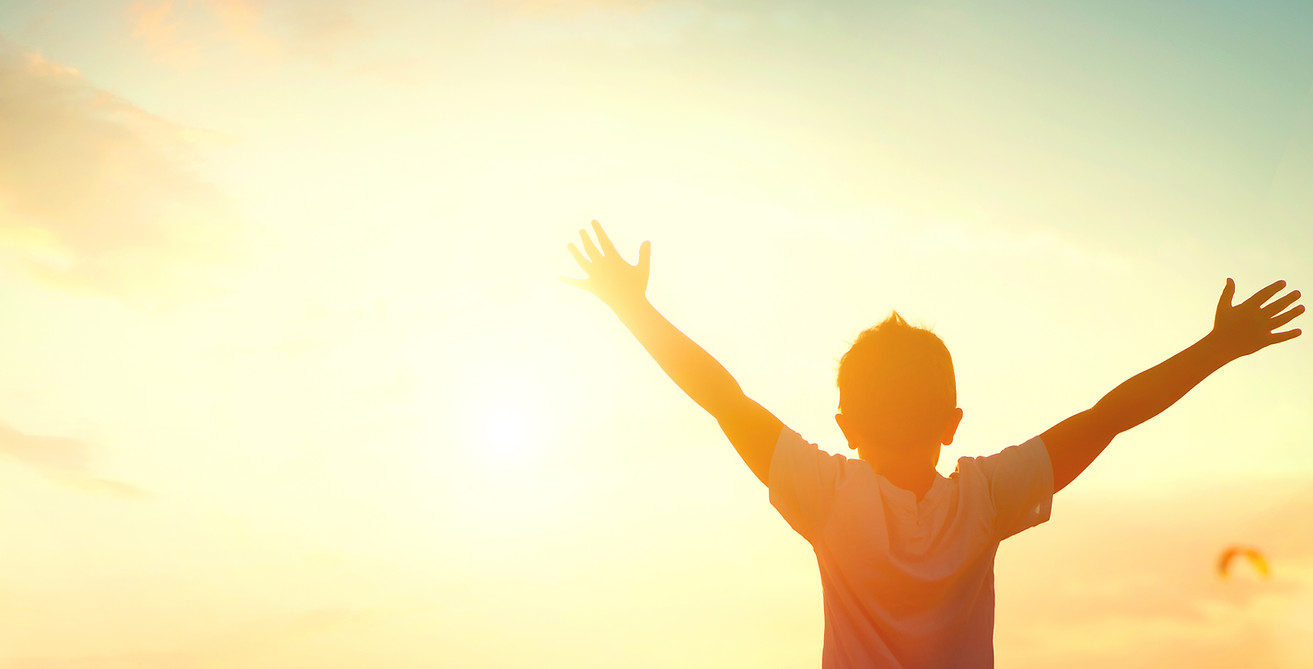 Little boy raising hands over sunset sky