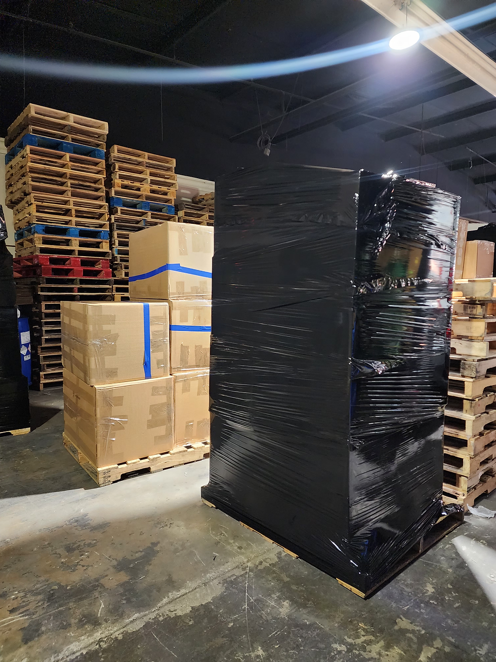 Large black plastic-wrapped pallet in a warehouse with stacked boxes and pallets.