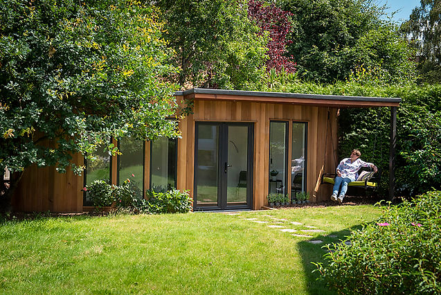 A beautiful Cedar Living Garden Room with cedar cladding nestled amoung oak trees with a lady sitting on the swing seat enjoying the sunshine.