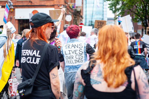 A shot taken in the middle of a march. Two people are out of focus in the foreground, back facing the camera. In front of them in focus is a person wearing a jacket that says "trans lives over cis feelings"