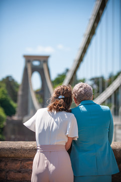 Two women, one in a blue suit, the other in a white and pink pantsuit, look off camera at the clifton suspension bridge, which is out of focus in the background