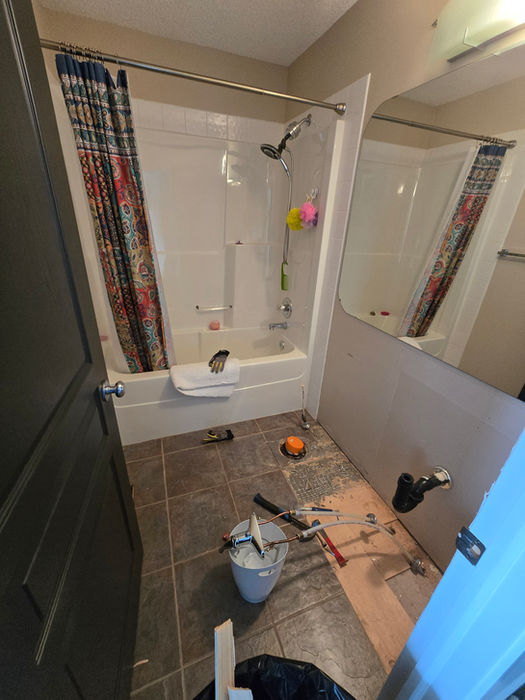 Before photo of a bathroom renovation showing beige walls, old vinyl flooring, a large wall-length mirror, and outdated lighting. The vanity has been removed, exposing the subfloor and plumbing, with tools and materials present in preparation for the renovation.