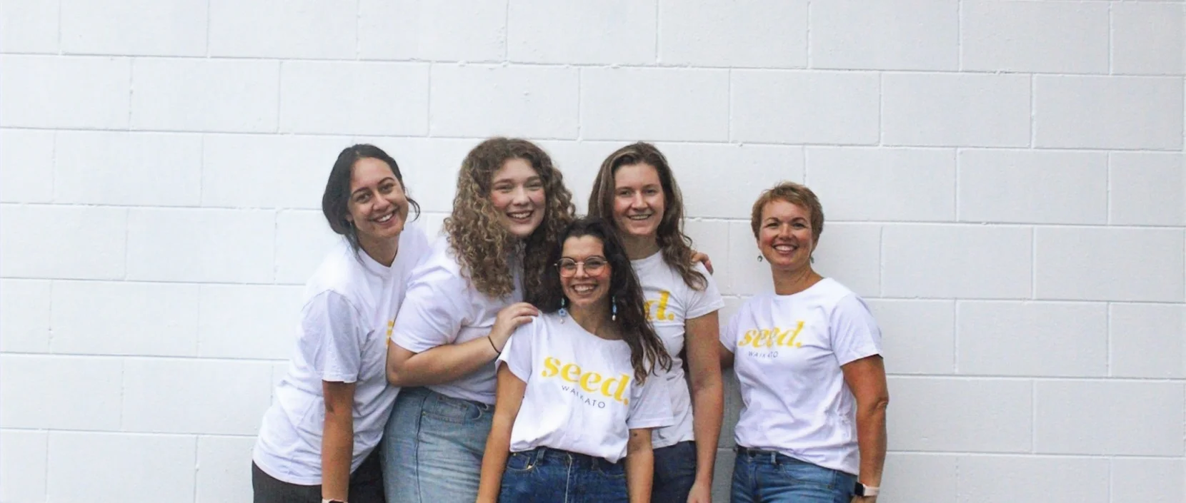 5 women in white tshirts with a a yellow logo reading Seed Waikato stand huddled in front of a white wall with big smiles on their faces. This is the Seeds for Change team for 2023