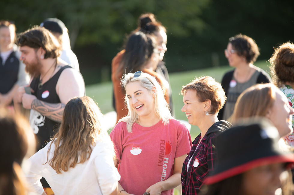 Three women stand in a circle laughing and talking together. You can see the faces of two women. One with blonde shoulder length hair wearing a pink shirt, and another with short darker hair wearing a black hoodie. The person facing away from the camera has long dark hair and is wearing a white shirt.