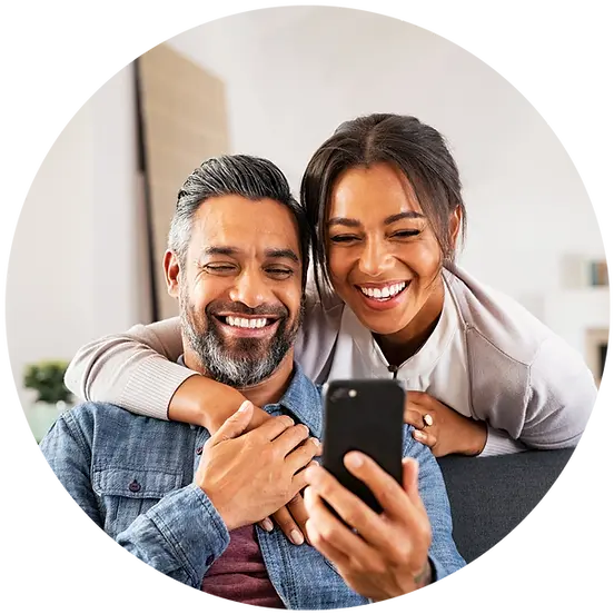 An man sitting on a couch smiling and looking at his phone with an woman behind him smiling and looking at the man's phone while embracing him from behind.