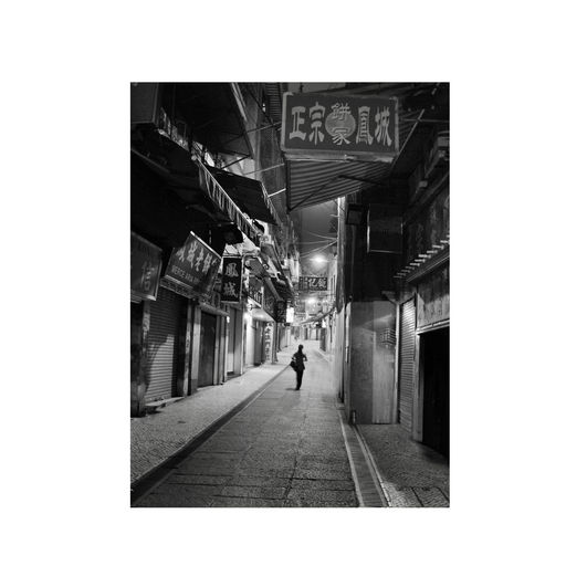 A candid black and white photograph of a girl walking late at night in Macau in a moody, noir-style nighttime city street, neon-lit streets, bokeh lights with signs and closed up shops.
