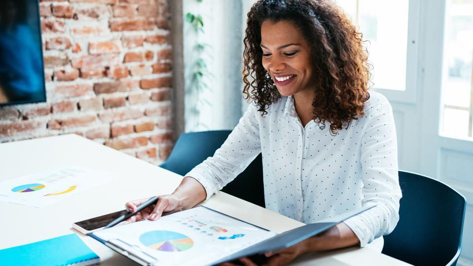 Femme souriante assise à un bureau, consultant un graphique coloré dans un dossier, avec un mur de briques en arrière-plan.