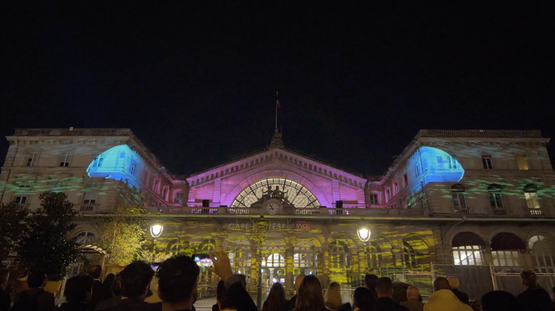 Projection lumineuse multicolore sur une façade historique, mettant en valeur ses détails architecturaux avec des teintes bleues et vertes, admirée par une foule enthousiaste