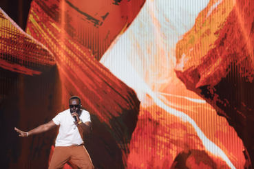 Un artiste de rap (GIMS) en pleine prestation, avec des visuels projetés en arrière-plan représentant un paysage numérique vibrant, créant une atmosphère immersive et rythmée.