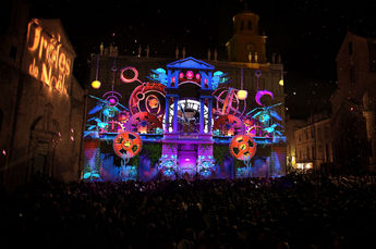 Une projection vibrante et colorée illumine un bâtiment avec des éléments de Noël, des jouets et des objets mécaniques, captivant le public massé sur la place, enchanté par ce conte visuel.