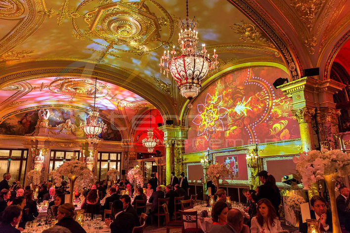Vue immersive d’une salle de réception somptueuse avec un plafond orné et des chandeliers, illuminée par des projections artistiques colorées, créant une atmosphère féerique inspirée de l’Art baroque.