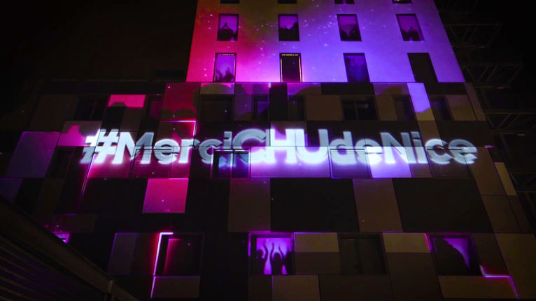 La façade du CHU de Nice illuminée avec le message "#MerciCHUdeNice" projeté en grand, symbolisant la reconnaissance envers le personnel hospitalier pour leur dévouement durant la pandémie. Des effets lumineux roses et violets renforcent l'impact visuel de cet hommage.