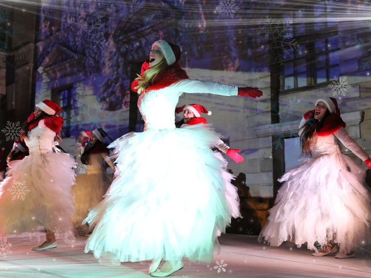 Des danseuses vêtues de tenues blanches et rouges dansent devant la façade illuminée. Leurs mouvements sont accompagnés de projections de flocons de neige et de décors hivernaux, plongeant le public dans une ambiance festive et magique propre à Noël.