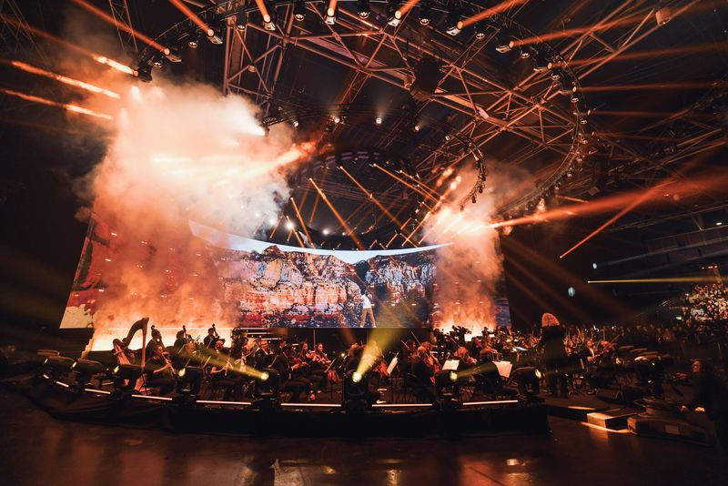 Concert spectaculaire de Gims avec des effets lumineux impressionnants, une grande scène entourée de musiciens et une ambiance immersive grâce à des projections visuelles et de la fumée.