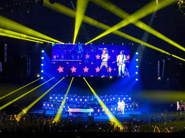 Vue de la scène avec Mika et un autre artiste interprétant une chanson devant une foule enthousiaste, tandis que des projections d’étoiles et des jeux de lumières jaunes illuminent l’arène.
