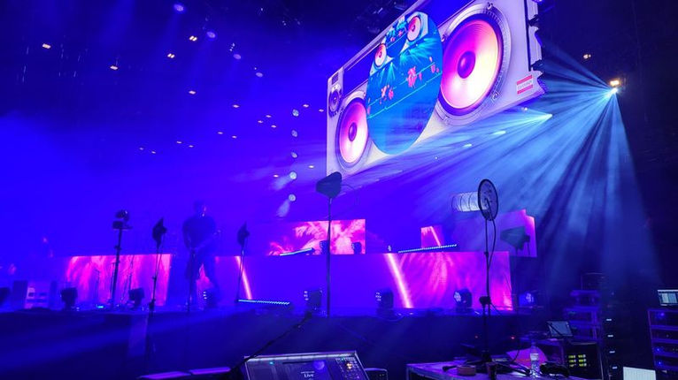 La scène est illuminée avec des tons violets et des animations visuelles sur des écrans géants représentant de grandes enceintes, créant une ambiance festive et dynamique pour l’événement spécial TF1 avec Mika.