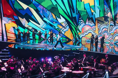 Une grande scène colorée avec un groupe de danseurs exécutant une chorégraphie devant des projections artistiques vibrantes, ajoutant de la profondeur et du dynamisme à la prestation. - Mika