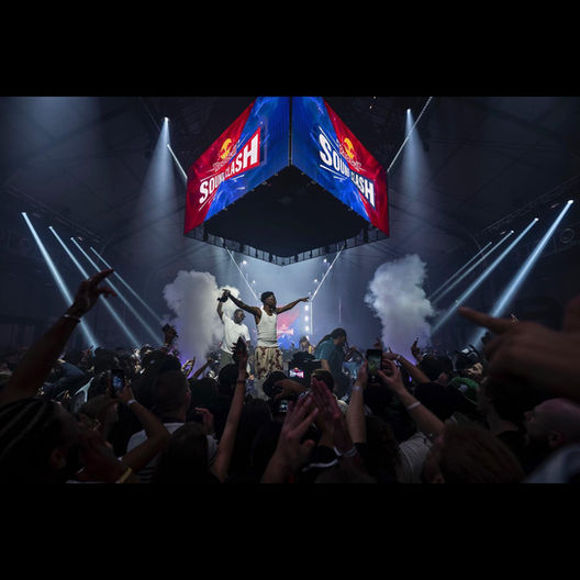 Un artiste se produit devant une foule en délire, entouré de faisceaux lumineux et d'effets de fumée, tandis qu'un grand écran suspendu affiche le logo du RedBull Soundclash. La scénographie numérique conçue par Studio ENORME accentue l’intensité de l’affrontement musical entre les artistes.