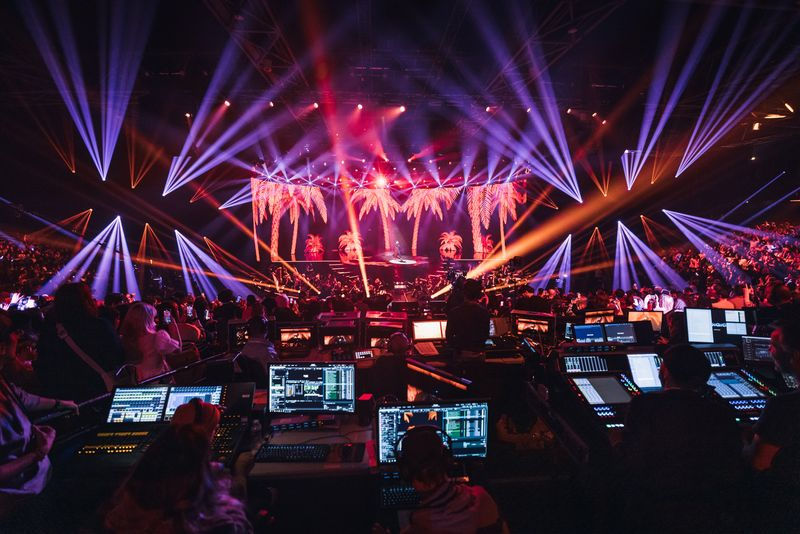 Vue d'ensemble de la régie technique pendant un spectacle en direct. L'équipe de production est entourée d'écrans et de consoles de contrôle, assurant la gestion des lumières et des visuels projetés. Des faisceaux lumineux bleus et violets illuminent la scène, créant une atmosphère immersive et spectaculaire pour le public. Les palmiers projetés à l'arrière-plan ajoutent une touche exotique à la scénographie.