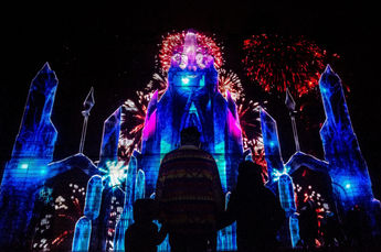 Une scène époustouflante projette un palais de glace entouré de feux d’artifice, plongeant les spectateurs dans un voyage à travers un monde de glace et de magie. Les silhouettes des spectateurs se détachent devant l’édifice illuminé.