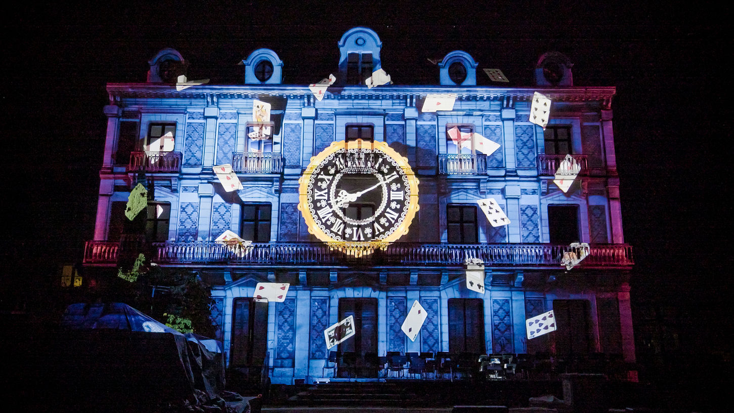 Mapping lumineux sur la façade de la Maison de la Magie, représentant une horloge centrale entourée de cartes à jouer en lévitation, créant une ambiance enchantée et mystérieuse.