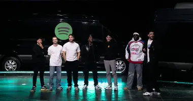 Les membres des deux équipes, rappeurs et streamers, se tiennent côte à côte sur scène, prêts pour la prochaine manche du concours. La scène est illuminée par des lumières vives et décorée par des visuels liés à Spotify.