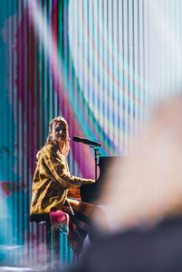 Un artiste (Angèle) jouant du piano sur scène, avec des projections visuelles créant un fond lumineux et abstrait, apportant une ambiance harmonieuse à sa performance lors d'un spectacle.