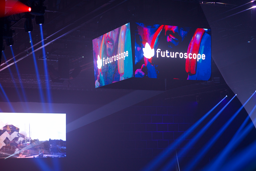 Un grand écran suspendu affiche le logo du Futuroscope, tandis que des faisceaux lumineux bleus envahissent la salle. Studio ÉNORME a conçu une scénographie visuelle immersive pour l'inauguration.