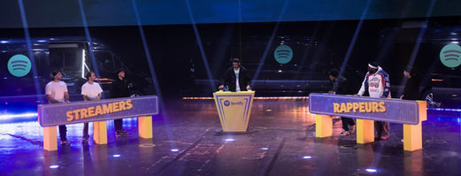 Les équipes de "Rappeurs" et de "Streamers" s’affrontent autour de pupitres marqués de leurs noms respectifs. L’atmosphère est compétitive, avec un éclairage de scène dramatique accentuant le côté festif de l’événement.