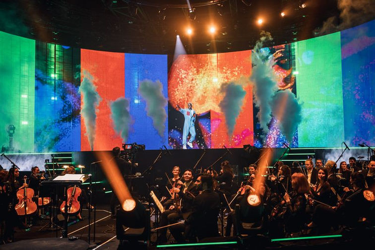 Un artiste chantant au centre d'une scène, entouré de projections numériques grandioses, tandis qu'un orchestre symphonique joue en arrière-plan, créant un mélange harmonieux entre musique live et visuels numériques.