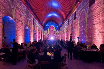 Vue de la salle remplie d’invités, entourés de projections colorées et historiques qui ornent les murs de la salle du Grand Tinel.