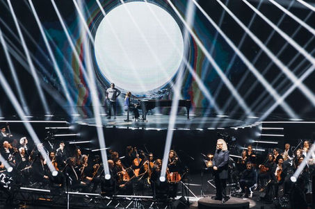 Un orchestre symphonique et Angèle se produisent devant un décor scénique spectaculaire, avec des effets lumineux qui mettent en valeur l'ampleur de la scène et la profondeur de l’événement.