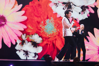 Un chanteur (Mika) en pleine performance, avec des fleurs géantes et colorées projetées en arrière-plan, créant une scène joyeuse et visuellement accrocheuse, renforçant le côté poétique de la chanson.