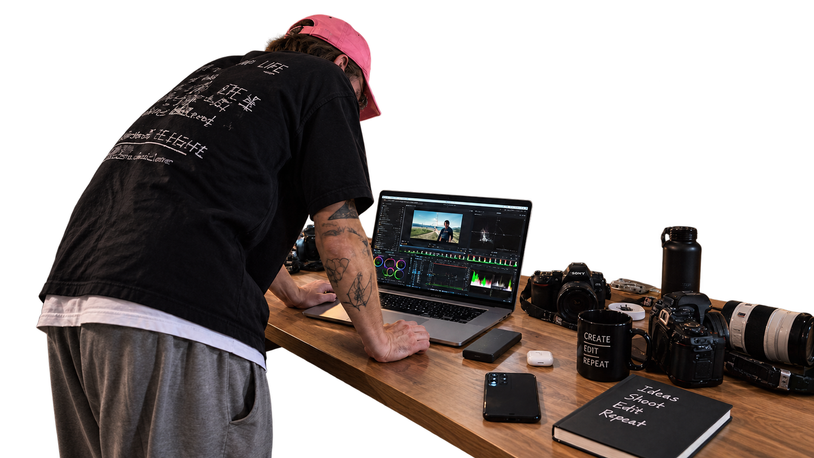 A person in a pink cap grading color at a wooden desk with a laptop and cameras.