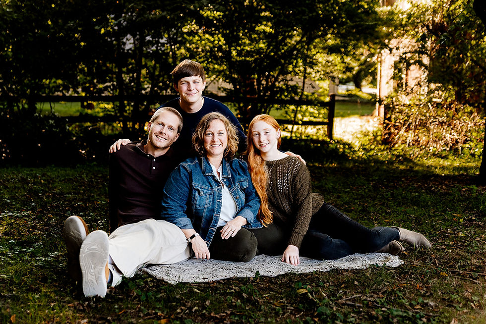 Family of four sitting on a blanket in a sunlit garden, smiling. Green foliage in the background, casual attire, and relaxed mood.