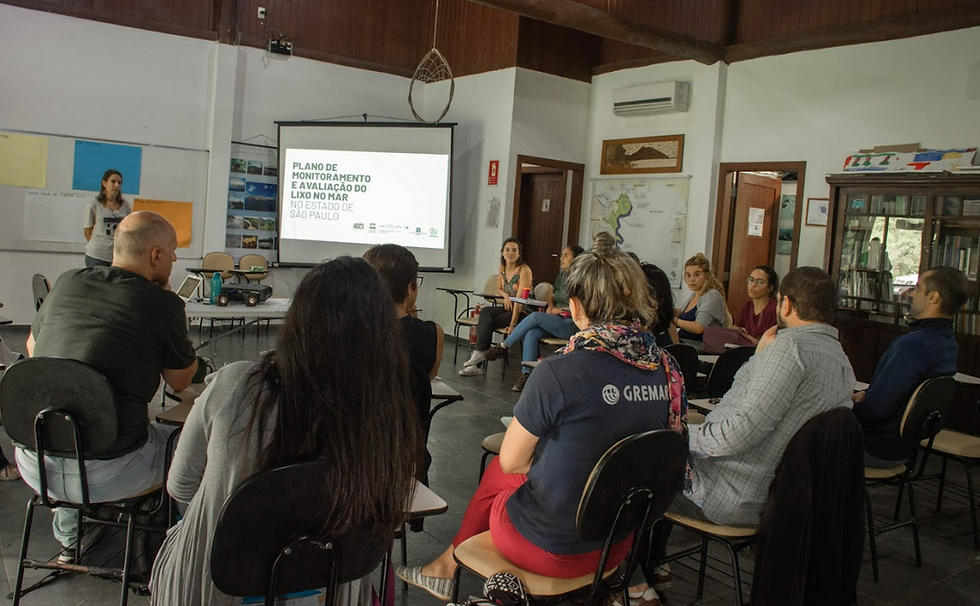 Fotografia de um grupo de 13 pessoas sentadas em cadeiras, organizadas em um semicírculo. As pessoas estão voltadas para uma tela, onde estão projetadas as palavras “Plano de Monitoramento e Avaliação do Lixo no Mar no Estado de São Paulo”. A sala tem paredes brancas, piso cinza escuro, uma lousa com cartazes coloridos, duas portas, um mapa do litoral de São Paulo e uma estante de livros