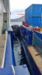 Photograph of two vessels side by side in the Arctic. Two large blue containers and several orange barrels are seen on the vessel on the right, with a single person in the background. A large buoy with tires is protecting the ships from collision with one another. In the distance the water seems calm and there are brown hills.