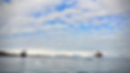 Photograph of an Arctic landscape. A blue sky with lots of white clouds takes up about two thirds of the frame. In the lower portion, to the left is the Maria S. Merian research vessel and to the right the Sonne research vessel. In the far background there are snow-capped mountains.