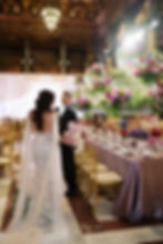 Bride in lace gown holding flowers walks with groom in elegant hall, with gold chairs, floral arrangements, and soft lighting creating romance.