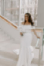 Bride in an off-shoulder white gown holding a bouquet, standing on a sleek staircase. Background shows large windows with trees.