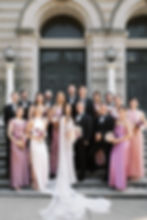 A wedding party in elegant attire poses on steps outside a historic building. Bridesmaids hold pink bouquets. The mood is joyful.