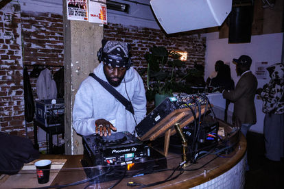 A tall black man who is wearing a grey hoodie, a black shoulder bag and a black bandana on his head. He is also djing.