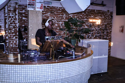 A black femme who is wearing white headphones, a black crop top, is wearing locs and is wearing a gold necklace and a few gold bracelets. She is djing.