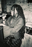 A greyscale photograph of a Black woman with long locs singing with a microphone in her hands. She is standing sideways and there is a brick wall behind her.